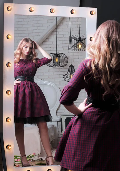 Reflected in large mirror with light bulbs, beautiful young girl with long purple plaid dress, holding her head in thought, looks into mirror. View from behind shoulder.