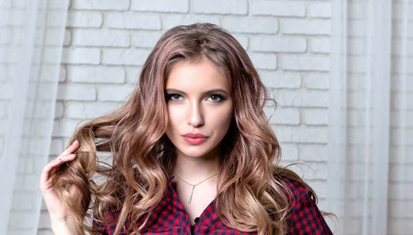 Portrait of young woman with long hair holding curl in her hand, looking intently at camera. Girl in red plaid shirt against white brick wall.