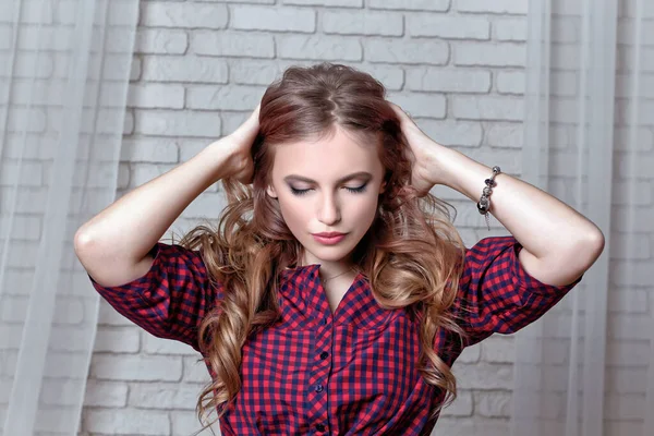 Beautiful young girl with long curls of hair with her eyes closed holds her hands behind her head against background of white brick wall.