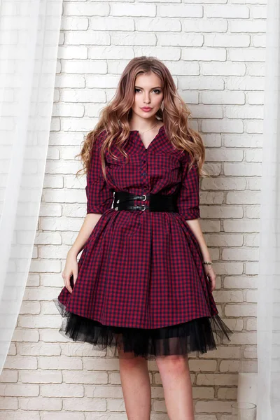 Beautiful young woman of model appearance with long brown hair in plaid dress stands against white brick wall, looking at camera.