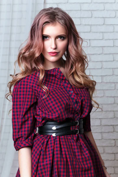 Young girl with long hair with curls on her shoulders, in plaid dress with black leather belt, stands against background of gray brick wall with curtain, looking into camera, medium plan, white loft room.