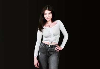 Beyaz bluzlu, ince, genç esmer, geniş göğüslü, siyah kot pantolonlu, bir eli belinde, kameraya bakıp gülümsüyor, siyah arka planda poz veriyor. Pankart, metin için yer.