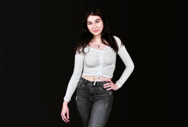 Beyaz bluzlu, ince, genç esmer, geniş göğüslü, siyah kot pantolonlu, bir eli belinde, kameraya bakıp gülümsüyor, siyah arka planda poz veriyor. Pankart, metin için yer.