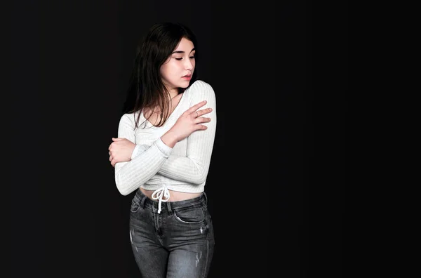 Slender young brunette in white blouse and black denim trousers crossed her arms over her chest, standing and looking at her shoulder against black background. Banner, place for text.