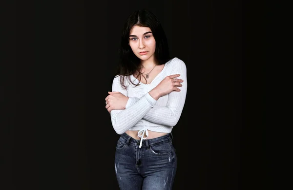Beyaz bluz ve siyah kot pantolon giyen çekici, kahverengi gözlü esmer kadın kollarını göğsünde kavuşturdu ve siyah arka plandaki kameraya baktı. Pankart, metin için yer.