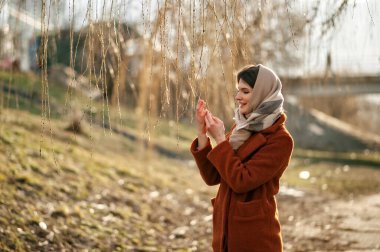 Paltolu ve atkılı genç esmer kız, güneşli bir günde, sonbahar parkının arka planında uzanan dalların olduğu ellerine bakar..