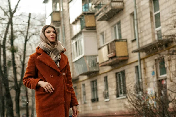 Kafasında fular olan turuncu ceketli genç bir kız kar altında dikilirken sağa bakıyor, çok katlı binanın zeminine karşı, fotokopi alanı ile..