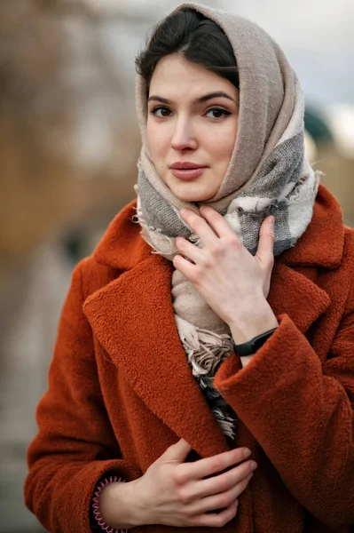 Genç esmer kızın portresi, büyük dudaklı, turuncu ceketli ve kafasında atkı olan, elleriyle yün eşarbı tutan, kameraya bakan, sonbahar parkının arka planındaki bulanık.