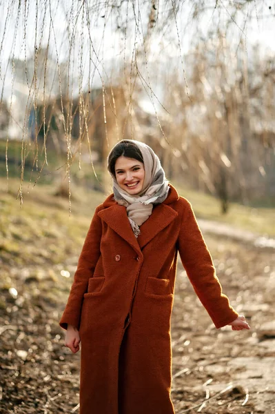 Turuncu ceketli, başörtüsü ve eşarplı, kollarını açmış, güçlü bir şekilde gülümseyen, kameraya bakan, ağacın dallarının altında duran, kopyalama alanı, sonbahar sezonu.