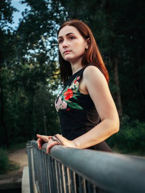 Siyah elbiseli, kahverengi saçlı, göğsünde çiçek desenleri olan genç bir kız. Köprüde duruyor. Korkuluklara yaslanıyor, yeşil ağaçların arkasına bakıyor..