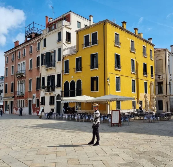  Venedik, İtalya, çizgili tişörtlü bir adam meydanın ortasında sarı bir evin arka planına doğru yürüyor.