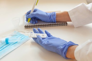 Sars covid antijen testi. Masada maske ve gözlük var. Doktor mavi eldivenleri elinde hızlı test ve yazma sonuçları tutuyor.