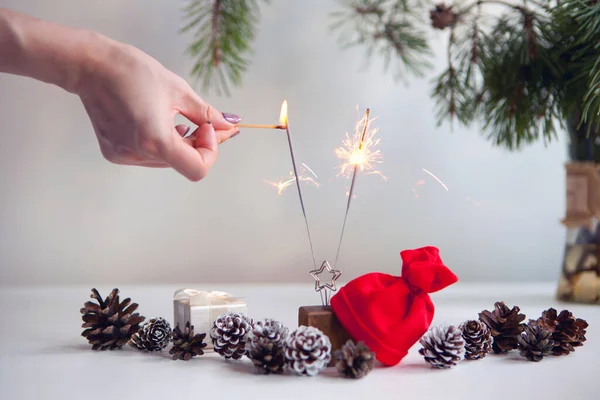 Mulher mão iluminação faíscas festivas perto de cones de pinho, árvore de natal e chapéu vermelho. Fogos de artifício partido Ano Novo — Fotografia de Stock