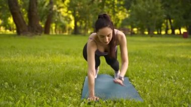 Parktaki bir kız spor için arka planda mavi bir spor paspasın üzerinde mor bir bluzla spor yapmaya gidiyor. Bir çayır ve ağaçlar.