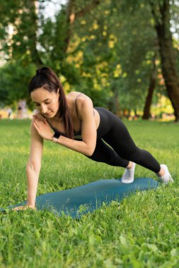 Parktaki bir kız spor için arka planda mavi bir spor paspasın üzerinde mor bir bluzla spor yapmaya gidiyor. Bir çayır ve ağaçlar.