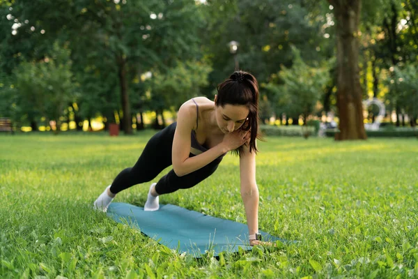 Parktaki bir kız spor için arka planda mavi bir spor paspasın üzerinde mor bir bluzla spor yapmaya gidiyor. Bir çayır ve ağaçlar.