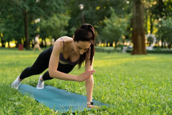 Parktaki bir kız spor için arka planda mavi bir spor paspasın üzerinde mor bir bluzla spor yapmaya gidiyor. Bir çayır ve ağaçlar.