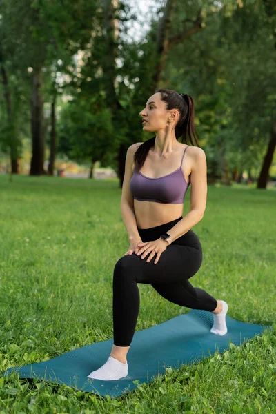 Parktaki bir kız spor için arka planda mavi bir spor paspasın üzerinde mor bir bluzla spor yapmaya gidiyor. Bir çayır ve ağaçlar.