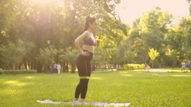 Parktaki bir kız spor için arka planda mavi bir spor paspasın üzerinde mor bir bluzla spor yapmaya gidiyor. Bir çayır ve ağaçlar.