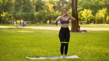 Parktaki bir kız spor için arka planda mavi bir spor paspasın üzerinde mor bir bluzla spor yapmaya gidiyor. Bir çayır ve ağaçlar.