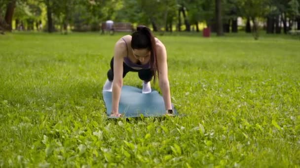 公園の屋外の女の子はスポーツのために行きます青のスポーツマットの上に黒のレギンスのスポーツ紫の上背景の牧草地と木 — ストック動画
