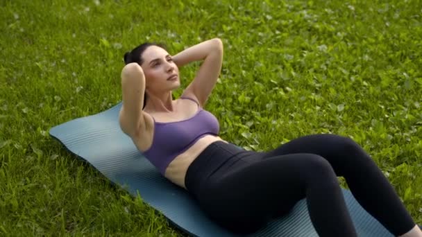公园里的一个女孩穿着紫色的运动上衣 穿着黑色的腿 站在蓝色的运动垫上 背景是草地和树木 — 图库视频影像