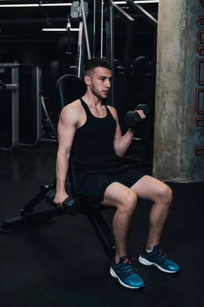 Adam spor salonundaki bir bankta otururken pazı için halterleri kaldırıyor. koyu arkaplan
