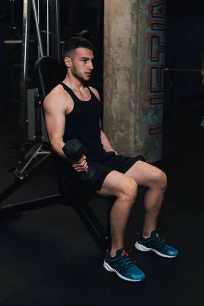 Adam spor salonundaki bir bankta otururken pazı için halterleri kaldırıyor. koyu arkaplan