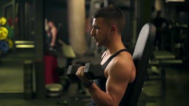 Adam spor salonundaki bir bankta otururken pazı için halterleri kaldırıyor. koyu arkaplan