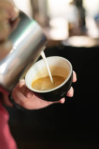 Kırmızı tişörtlü barista siyah bardağa kahve doldurur.