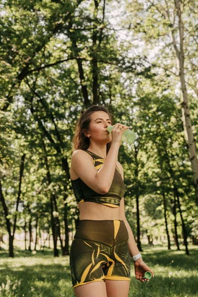 young girl drinks water in the park, in the forest. in a green tracksuit