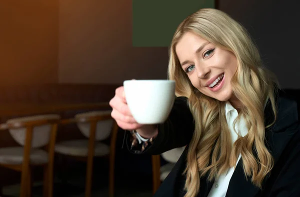 Mujer rubia joven sosteniendo taza con café y sonriendo a la derecha en la cámara. Feliz tiempo de descanso en el trabajo. Café concepto comercial con adorable y hermosa joven. — Foto de Stock