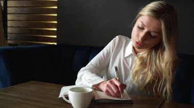 Gorgeous girl make business notes with pen on coffee break in restaurant. Startup office worker drinking coffee. Young woman smiling while working. High quality 4k footage