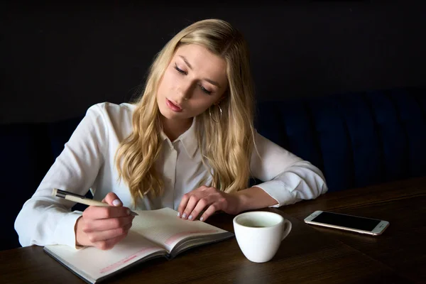 Beautiful young woman work in caffe. Woman writing and using phone while sitting in restaurant. High quality photo