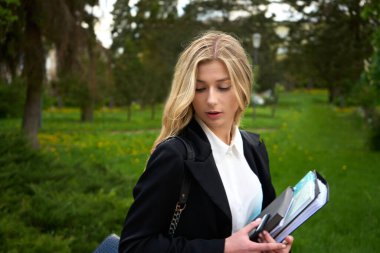 Resmi iş elbiseleriyle parkta gezen ve açık yaz gününde doküman okuyan sarışın genç bir bayan. Güzel, modern iş kadını konsepti. Yüksek kalite fotoğraf