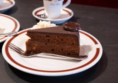 Avusturya kaynaklı kayısı reçelli Sachertorte çikolatalı pastasından bir parça. Sütlü beyaz tahtada krema ve siyah kahve ile servis ediliyor.