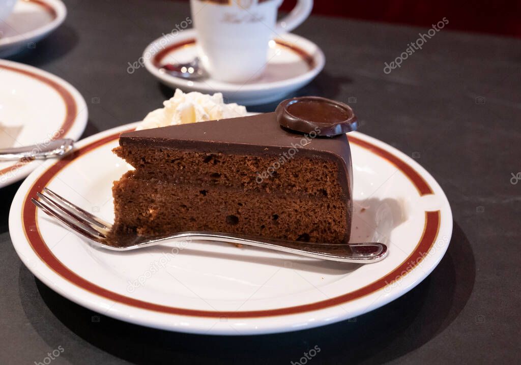 Piece of famous Sachertorte chocolate cake with apricot jam of Austrian origin served with whipped cream on white board with black coffee