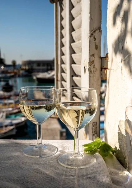 Cassis şaraplarından elde edilen iki bardak beyaz şarap, Fransa, Provence, Cassis 'te renkli tekneli eski balıkçıların limanı manzaralı terasta servis edilir.