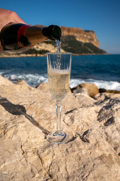 Pouring of French champagne sparkling wine in glass and view on beach, blue sea water and mountains in Cassis, Provence, France