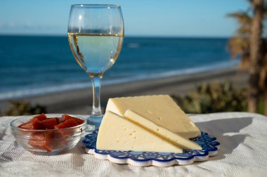 İspanyol sert keçi peyniri, açık havada membrillo reçeli ve arka planda mavi deniz bulunan bir bardak beyaz şarapla servis edilir.