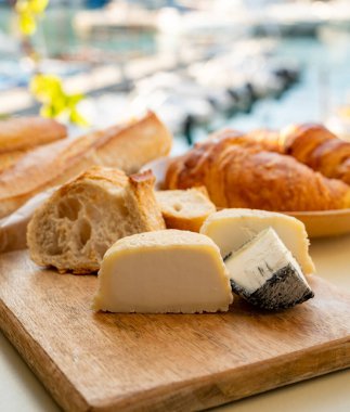 Provence 'de güneşli bir sabah, taze kruvasan, baquett ekmeği, crottin keçi peyniri ve Cassis, Provence, Fransa limanındaki balıkçı tekneleri manzaralı bir kahvaltı.