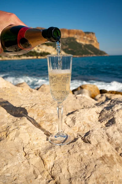 Pouring of French champagne sparkling wine in glass and view on beach, blue sea water and mountains in Cassis, Provence, France