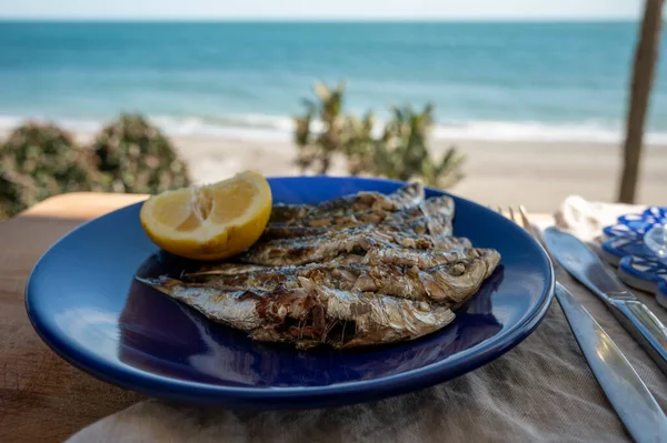 Sardines espeto prepared on skewers and open flame on fireplace with olive trees wood, served outdoor with and view on blue sea, Malaga, Andalusia, summer vacation in Spain