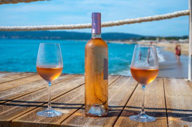 Provence 'den getirilen bardaklar ve bir şişe soğuk gül şarabı mavi deniz manzaralı ahşap yat iskelesinde ve Saint-Tropez yakınlarındaki plaj plajı Plage de Pampelonne, Fransa' da yaz tatili
