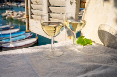 Cassis şaraplarından elde edilen iki bardak beyaz şarap, Fransa, Provence, Cassis 'te renkli tekneli eski balıkçıların limanı manzaralı terasta servis edilir.