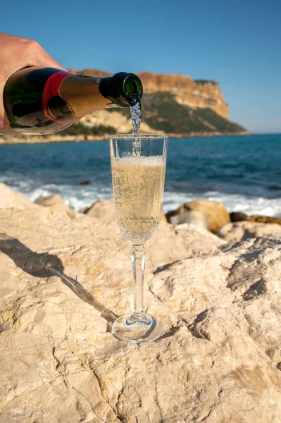 Pouring of French champagne sparkling wine in glass and view on beach, blue sea water and mountains in Cassis, Provence, France