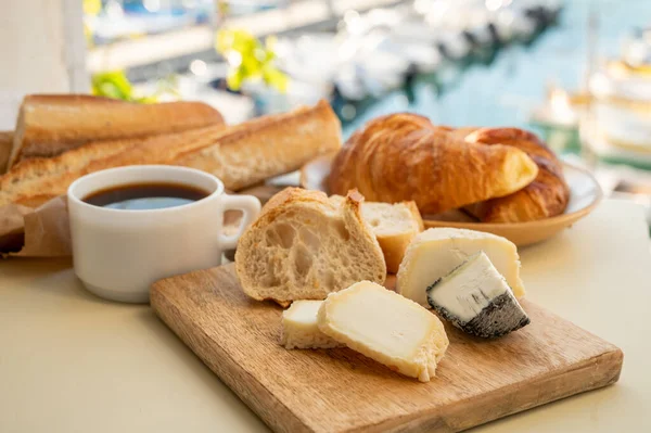 French breakfast with fresh baked croissants, baquett bread, crottin goat cheese, black coffee and view on fisherman\'s boats in harbour of Cassis, Provence, France. Vacation in Provence.
