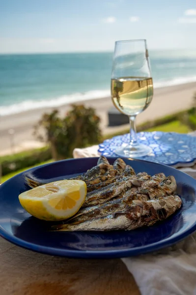 Sardines espeto prepared on skewers and open flame on fireplace with olive trees wood, served outdoor with lemon and glass of fino sherry wine and view on blue sea, Malaga, Andalusia, summer vacation in Spain