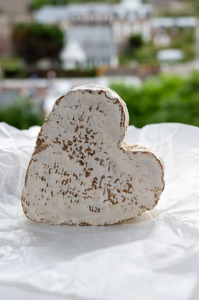 French heartshaped whole piece of neufchatel cow cheese on white paper and view on old houses of Etretat, Normandy, France