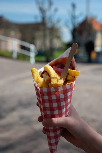 Dutch and Belgian fast and street food, child\'s hand with paper bag of fried potatoes chips with tomato ketchup